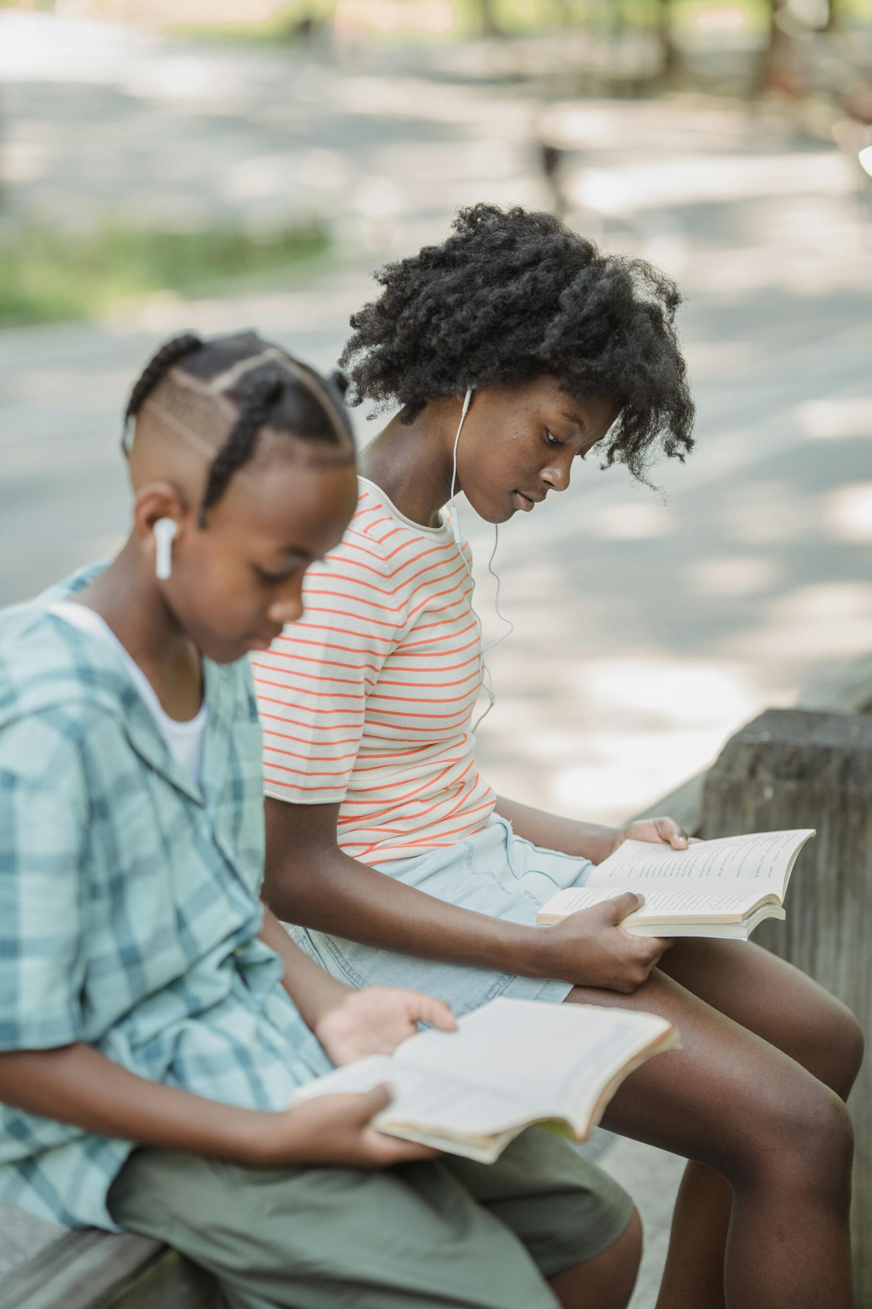 Two teenagers sitting on a bench outdoors, engrossed in books, with earphones on.